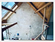 Innenraum mit hoher Decke aus hellen Ziegelsteinen und sichtbaren Holzbalken sowie Dachkonstruktion. Unten sind große Fensterflächen und eine moderne Hängelampe zu sehen, mit etwas Wandmontage und Ablage rechts.