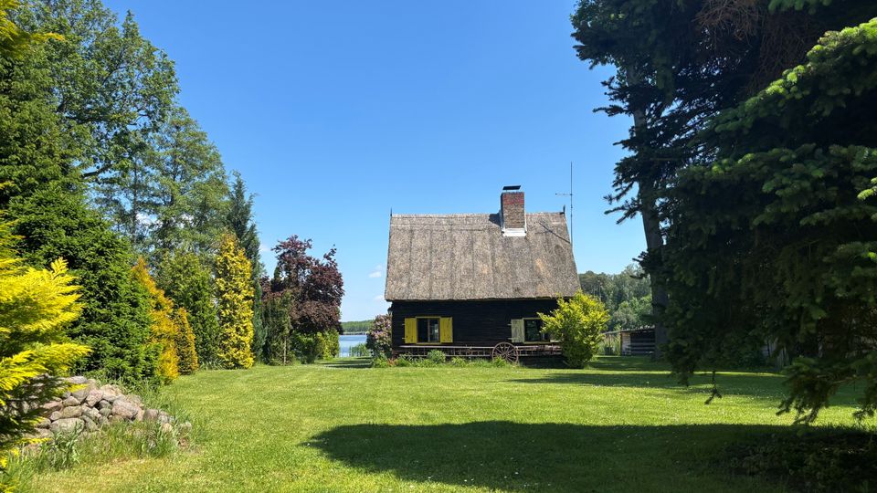 Kleines Holzhaus mit Reetdach und Schornstein am Ende einer großen Rasenfläche, eingerahmt von hohen Bäumen und Sträuchern. Im Hintergrund ist zwischen den Bäumen ein See zu sehen.