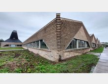 Ein modernes Gebäude mit markanter Klinkerfassade und gezacktem Dachdesign, ergänzt durch große Glasfenster. Im Hintergrund ist ein weiteres Gebäude mit einer schwarzen, pyramidenförmigen Dachkonstruktion sichtbar, umgeben von einer Grünfläche und Gehwegen.