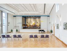 Innenansicht eines Veranstaltungsraumes mit einer Bühne, auf der ein großformatiges Wandbild eines Flugzeugs zu sehen ist. Der Raum ist mit Reihen von Tischen und Stühlen ausgestattet, hat helle Wände, Parkettboden und große Fenster; im Vordergrund steht ein schwarzer Flügel.