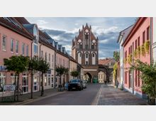 Straßenszene in einer historischen Altstadt mit gepflasterter Fahrbahn, flankiert von pastellfarbenen Gebäuden. Im Hintergrund ein gotisches Stadttor aus Backstein mit dekorativen, spitzbogigen Elementen und einer Durchfahrt.