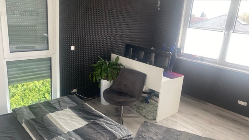 Modernes Schlafzimmer mit dunkler, strukturierter Wandverkleidung, einem Bett mit grauer Bettwäsche und hellem Laminatboden. Daneben stehen ein weißer Schreibtisch mit Monitoren, ein grauer Stuhl, eine Zimmerpflanze sowie große Fenster und eine Glastür mit Rollläden.