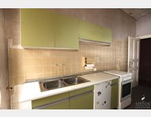 Alte Küche mit pastellgrünen Schränken, beige gefliester Rückwand und einer Edelstahl-Spüle mit zwei Becken. Rechts befinden sich ein kleines weißes Herd- und Ofenmodul sowie ein geöffneter Holztürrahmen.