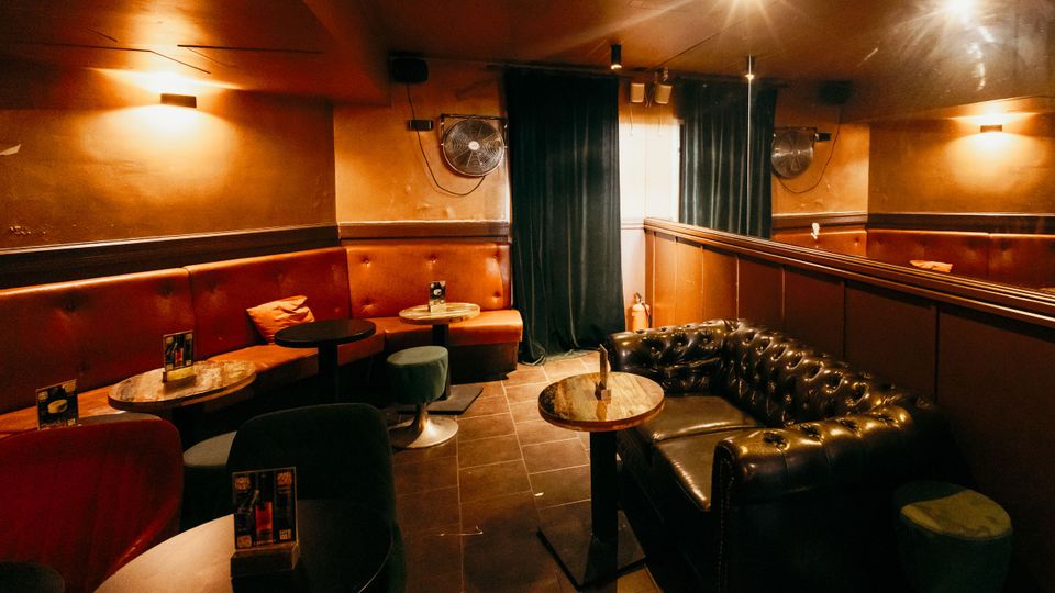 Gemütlicher Lounge- oder Bar-Innenraum mit gepolsterten Sitzbänken in Rot und einem schwarzen Chesterfield-Sofa, runden Marmortischen und dunklem Fliesenboden. Warme Wand- und Deckenbeleuchtung, Spiegelwand sowie Vorhänge und Wandventilatoren prägen den Raum.