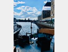 Ein Blick von einem Boot auf einen Fluss mit zwei schwarzen Sesseln und einem kleinen Tisch im Vordergrund. Im Hintergrund sind die Oberbaumbrücke, moderne Gebäude am Ufer und ein teils bewölkter Himmel zu sehen.