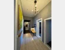 Ein schmaler Flur mit hellen Holzdielen und grauen Wänden, dekoriert mit einem Kronleuchter und einem großformatigen Gemälde an der linken Wand. Rechts befinden sich mehrere offene Türrahmen, eine Holzbank mit einer Tischlampe steht an der Wand, und im Hintergrund ist ein weiterführender Raum sichtbar.