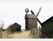 Eine historische Windmühle aus Holz mit Flügeln und einem kleinen Windrad steht auf einem ländlichen Gelände, umgeben von weiteren Holzbauten, einem Zaun und kahlen Bäumen. Die Umgebung wirkt wie eine weitläufige Wiesen- und Felderlandschaft.