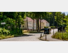 Eine gepflasterte Straße führt durch eine von Bäumen gesäumte Umgebung zu mehreren zweigeschossigen, gelb gestrichenen Gebäuden mit roten Ziegeldächern. Im Vordergrund steht ein Straßenschild mit der Aufschrift „Landeskriminalamt“.