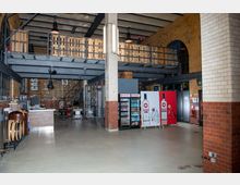 Industrieähnlicher Verkaufsraum mit Backsteinwänden, Metallträgern und offenem Zwischengeschoss. Im Vordergrund befinden sich Verkaufsregale mit Getränken und Werbetafeln sowie eine Theke, im Hintergrund führen Treppen zu einer oberen Ebene.