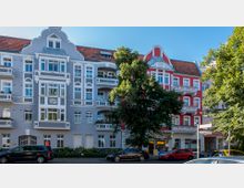 Straßenszene mit zwei nebeneinanderliegenden Altbauwohnungen, einem graublauen und einem roten Gebäude mit weißen Akzenten, verzierten Fassaden und Balkonen. Vor den Gebäuden stehen Autos, und ein großer Baum verdeckt teilweise die Ansicht.