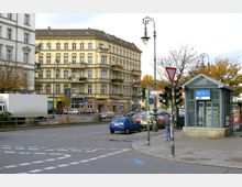 Straßenszene an einer Kreuzung mit historischen Wohngebäuden im Hintergrund, einer Glasstruktur für den U-Bahn-Zugang (U7) im Vordergrund und mehreren Verkehrsschildern, Autos und Fußgängern. Die Umgebung ist städtisch mit Bäumen am Straßenrand und herbstlichem Laub.