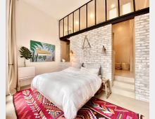 Schlafzimmer mit modernen und wohnlichen Elementen, featuring einer weißen Backsteinwand, einem Doppelbett mit weißer Bettwäsche und einem bunten Teppich mit geometrischem Muster. Im Hintergrund sind eine erhöhte offene Ebene mit Glaswänden und sichtbare Beleuchtung sowie eine dekorative Wandkunst und Pflanzen zu sehen.