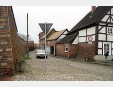 Eine schmale Kopfsteinpflasterstraße in einem historischen Dorf mit Fachwerkhäusern aus rotem Ziegel und weißem Putz. Links verläuft eine Backsteinmauer, rechts steht ein Verkehrsschild, ein geparktes Auto und ein Fahrrad an einem Haus.