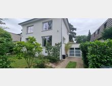 Zweistöckiges, weiß verputztes Wohnhaus mit großen Fenstern, umgeben von einem gepflegten Garten mit Bäumen und Sträuchern. Rechts im Bild ein gepflasterter Weg und eine angrenzende Garage mit modernen Glastüren.