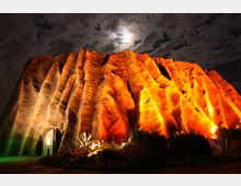 Beleuchtete Felsformation in einem Wüstengebiet, angestrahlt von warmen, orangefarbenen Lichtern, mit Pflanzen im Vordergrund und wolkigem Nachthimmel im Hintergrund. Ein kleiner Eingang mit einer beleuchteten Tür ist in die Felsen integriert.