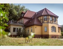 Gelbes zweistöckiges Fachwerkhaus mit roten Ziegeldächern, mehreren Fenstern und einer Terrasse mit Geländer; das Gebäude steht inmitten eines grasbewachsenen Gartens mit vereinzeltem Bewuchs und Bäumen im Hintergrund.