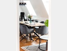 Esszimmer in einem modern eingerichteten Wohnraum mit Holzfußboden, einem rechteckigen Holztisch, gepolsterten, grauen Stühlen mit Rautenmuster und einer hängenden Lampe mit drei Glaskugeln. Im Hintergrund befinden sich Schrägfenster, ein Schreibtischstuhl und Dekoration, darunter eine Vase mit Blumen.