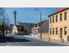Straßenszene in einer kleinen Stadt mit Wohnhäusern mit Satteldächern, einem gelben Mehrfamilienhaus und einem markanten Backsteinturm im Hintergrund. Gepflegte Gehwege, Bäume entlang der Straße und ein Schild mit der Aufschrift „Business Park“ sind sichtbar.