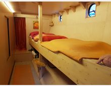 Ein schmaler Innenraum aus hellem Holz mit einem Hochbett, das orangefarbene und rote Decken hat. An der Wand befinden sich runde Fenster, ein paar kleine Regale und ein Vorhang in Rot, der vermutlich einen weiteren Bereich abtrennt.