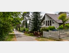 Schmale Wohnstraße mit Einfamilienhäusern, gesäumt von Bäumen und Gärten. Im Vordergrund ein weißes Haus mit Satteldach und weißem Zaun, im Hintergrund mehrere weitere Häuser und geparkte Autos.