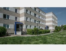 Mehrstöckiges Wohngebäude mit grauer Fassade und horizontalen Streifen aus hellem Putz. Vor dem Gebäude befinden sich grüne Büsche, Rasenflächen mit vereinzelt blühendem Löwenzahn sowie ein Gehweg und Laternen.