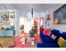Bright, eclectic living room with pale blue walls, a deep blue sectional sofa with colorful pillows, and a pink patterned rug around a glass coffee table. The space features a gallery wall of framed art above a low cabinet, a sculptural hand-shaped chair, hanging pendant lights, and double doors opening to an adjoining room.