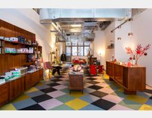 Innenraum eines modernen Ladengeschäfts mit Holzmöbeln, buntem Schachbrett-Bodenmuster und offener Deckenverkleidung mit sichtbaren Lüftungsschächten. Auf Regalen und Tischen sind verschiedene Produkte wie Bücher, Textilien und Dekorationsartikel ausgestellt, während im Hintergrund ein großes Fenster und Sitzmöbel zu sehen sind.