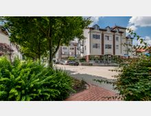 Straßenszene mit einem mehrstöckigen Gebäude im hellen Putzstil, das die Aufschrift "Kantine im Gutshof" trägt. Im Vordergrund befinden sich grüne Büsche und Bäume entlang eines Gehwegs, im Hintergrund ist ein kleiner Parkplatz mit mehreren geparkten Autos zu sehen.