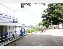 Ein Weg entlang eines Yacht- und Bootsverleihs an einem Seeufer mit Blick auf Boote im Wasser, umgeben von Bäumen und Schilf. Im Vordergrund steht ein Zaun mit Schildern und ein Automat, rechts Skulpturen und Fahrradständer.