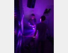 Zwei Personen befinden sich in einem kleinen Raum mit schräger Decke, beleuchtet von violettem und blauem Licht. An der Wand befinden sich schallabsorbierende Schaumstoffplatten, und oben ist ein Lautsprecher erkennbar.