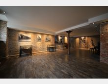 Innenraum einer Galerie oder Ausstellungshalle mit freiliegenden Backsteinwänden, mehreren gerahmten Bildern an der Wand, Holzfußboden und punktueller Deckenbeleuchtung. In der Mitte des Raumes befinden sich einzelne Glasvitrinen und ein metallener Stützpfeiler.