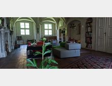 Wohnraum mit historischem Ambiente, gekennzeichnet durch gewölbte Decken, große Fenster und eine Mischung aus modernen und klassischen Möbeln. Im Vordergrund ein grüner Zimmerpflanze, daneben moderne Sofas, Bücherregale und ein Kamin aus Stein an der linken Wand.