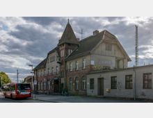 Historisches Bahnhofsgebäude mit markantem Turm und gemischter Backstein- und Verputzfassade, teils mit Graffiti versehen. Im Vordergrund steht ein roter Linienbus an einer Haltestelle, und der Himmel zeigt dichte Wolken.