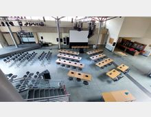 Ein moderner Veranstaltungsraum mit Reihen von Stühlen und Tischen, die mit Mikrofonen ausgestattet sind, sowie einer Bühne und einer großen Leinwand im Hintergrund. Im Raum befinden sich zudem Beleuchtungs- und Audiotechnik, eine Bar und ein Treppenaufgang im Vordergrund.