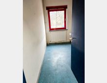 Schmales Zimmer mit blauen Bodenfliesen, weiß gestrichenen Wänden und einem roten Fensterrahmen. Unter dem Fenster befindet sich ein Heizkörper, rechts steht ein kleines Waschbecken, und die Türe ist dunkelblau.