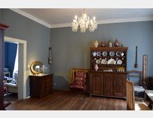 Historisches Wohnzimmer mit Holzboden, grauen Wänden und einem Kronleuchter an der Decke. Im Raum stehen ein antiker Schrank mit Geschirr und Keramik sowie weitere Möbel wie eine Kommode mit Tischlampe, ein Polsterstuhl und ein Esstischstuhl.