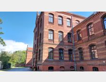 Backsteingebäude mit historischen Zierelementen und Segmentbogenfenstern, umgeben von Bäumen und einem gepflasterten Weg bei sonnigem Wetter. Im Hintergrund ist ein weiteres rotes Gebäude sowie Grünanlage sichtbar.