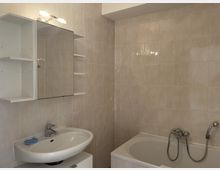 Beigefarbenes Badezimmer mit einer gefliesten Wand, einem Waschbecken unterhalb eines Spiegelschranks mit eingebauten Regalen und einer Badewanne mit Wandarmatur und Duschschlauch.