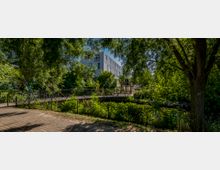 Ein kleiner Park mit dichtem Grün, einem Weg aus Pflastersteinen und einer schmalen Brücke über einen Bach. Im Hintergrund ist ein modernes, weißes Gebäude sichtbar.