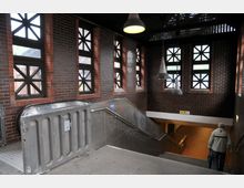 Treppenhaus eines U-Bahn- oder Bahnhofsgebäudes mit Backsteinwänden und Fenstern im Kreuzmuster. Sichtbar sind eine Rolltreppe, eine Treppe mit Geländer, Hängelampen und ein Mensch, der die Treppe hinuntergeht.