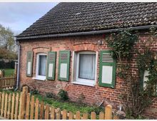 Kleines Backsteinhaus mit Satteldach und grün-weißen Fensterläden, umgeben von einem niedrigen Holzzaun. Vor dem Haus befinden sich ein kleiner Garten mit Blumen und Kletterrosen sowie ein bewölkter Himmel im Hintergrund.