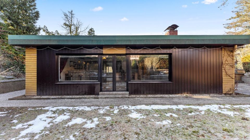 Flacher, eingeschossiger Bungalow mit dunkel vertäfelter Holzfassade, grünem Flachdach und mittiger Glastür zwischen zwei großen Fensterfronten. Davor liegt ein Garten mit fleckigem Schnee, umgeben von Bäumen und niedrigen Mauern.