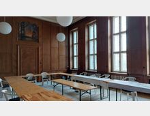 Holzvertäfelter Raum mit großen, hohen Fenstern, die viel Tageslicht hereinlassen. Mehrere hölzerne Tische und weiße Plastikstühle sind entlang der Wände angeordnet, darüber hängen runde Deckenleuchten.