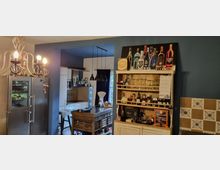 Küche mit modernem Design, bestehend aus einem hohen silbernen Kühlschrank, einem Holztisch, und einer weißen Einbauküche mit dunkler Herdplatte. Links ein Kronleuchter, rechts ein Holzregal mit Flaschen und Gläsern sowie einer großen dekorativen Abbildung von Getränkeflaschen. Im Hintergrund ist ein Fenster mit dunklen Vorhängen sichtbar.