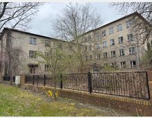 Mehrstöckiges, hellbraunes Gebäude mit verputzter Fassade und mehreren Fenstern, teilweise beschädigt. Vor dem Gebäude befindet sich eine schwarze Metallzaunanlage, bepflanzt mit Büschen und unbelaubten Bäumen; der Boden ist mit Gras und Moos bedeckt.