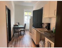 Schmale Küche mit weißer Schrankzeile und grauer Arbeitsplatte samt Spüle und Kochfeld; entlang der Wand steht ein kleiner Esstisch mit vier Stühlen. Am Ende des Raums befinden sich ein großes Fenster und ein Kühlschrank, der Boden ist in Holzoptik gehalten.