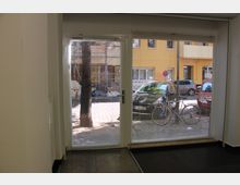 Blick aus einem Innenraum durch eine gläserne Tür mit Jalousien auf eine Straße mit parkenden Autos, Fahrrädern und einem gelben Gebäude im Hintergrund. Draußen sind Bäume und Bauarbeiten zu sehen.