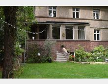 Zweistöckiges Wohnhaus mit Putz- und Ziegel-Fassade, eine kleine Treppe führt von einem Garten zu einer überdachten Terrasse. Der Garten ist gepflegt, mit Blumenbeeten, Rasen, einer Lichterkette und Bäumen im Hintergrund.