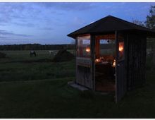 Kleiner hölzerner Pavillon mit geöffnetem Eingang und Fensterfolien, innen warm beleuchtet, auf einer Wiese am Rand eines weiten Feldes. Im Hintergrund stehen Bäume am Horizont und mehrere Pferde weiden auf der Koppel bei Abenddämmerung.