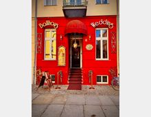 Fassade eines Gebäudes mit rotem Anstrich und dem Schriftzug „Ballhaus Wedding“. Im Mittelpunkt befindet sich ein Eingangsbereich mit rotem Vordach, einer Treppe mit rotem Teppich und flankierenden goldenen Absperrungen.